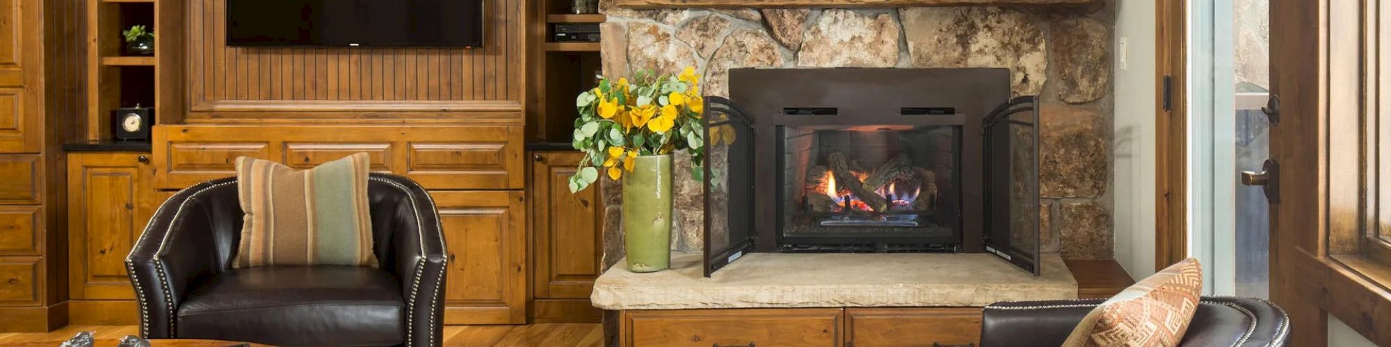 A cozy living room features a stone fireplace, two leather chairs, a wall-mounted TV, a colorful painting, and wooden cabinetry.