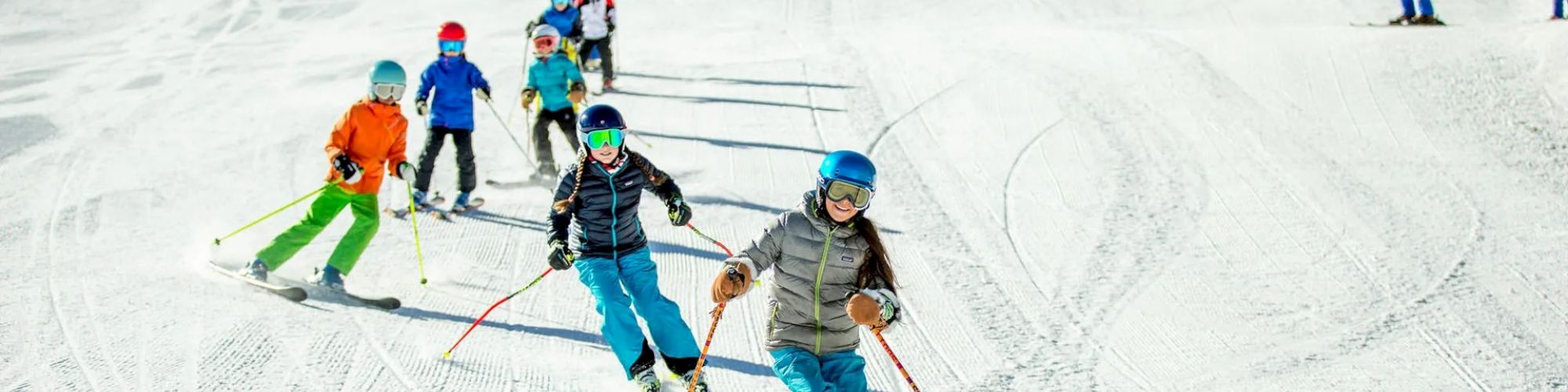 A group of children in colorful outfits are skiing down a snowy slope, with an instructor in blue leading them. The sky is clear and bright.