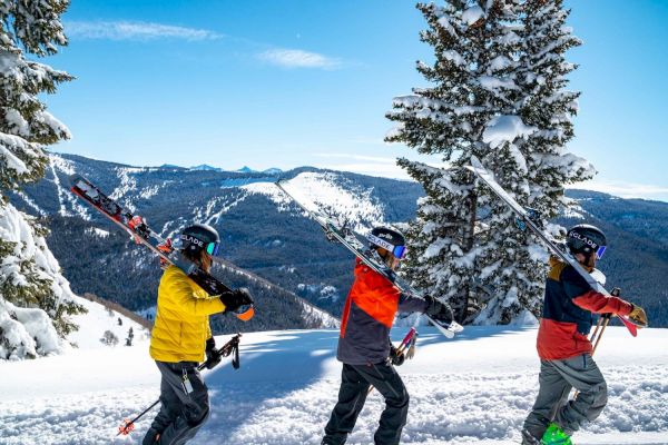 Three people carrying skis on their shoulders walk through a snowy landscape with mountains and trees in the background, under a clear blue sky.