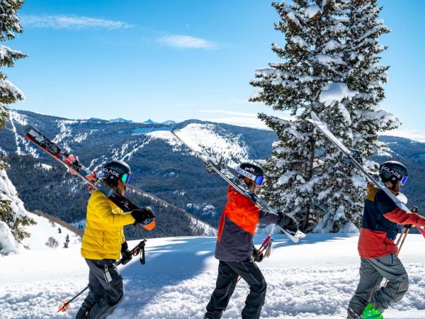 Three people carrying skis on their shoulders walk through a snowy landscape with mountains and trees in the background, under a clear blue sky.