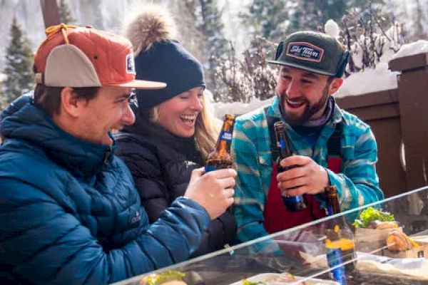 Three people are sitting outdoors in a snowy setting, smiling and clinking beer bottles, enjoying their time together.