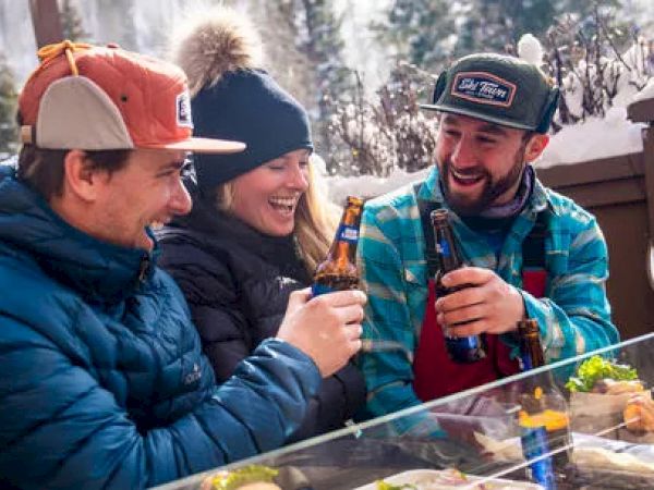 Three people are sitting outdoors in a snowy setting, smiling and clinking beer bottles, enjoying their time together.