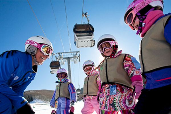 A group of children dressed in winter gear and helmets is being instructed by an adult on a snowy mountain with ski lifts in the background.