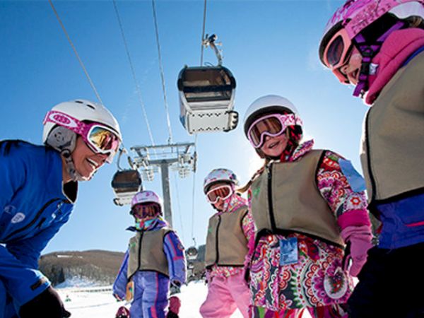 A group of children dressed in winter gear and helmets is being instructed by an adult on a snowy mountain with ski lifts in the background.