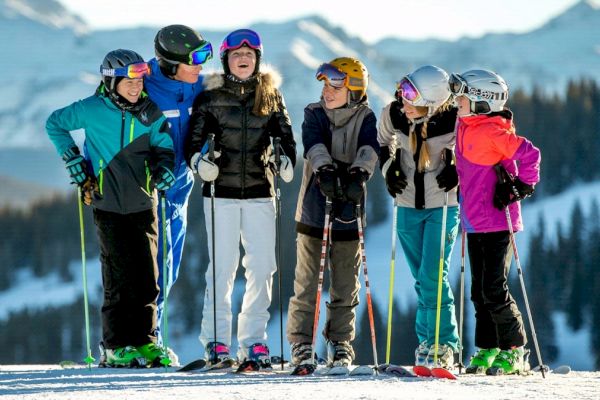 A group of six people in winter clothing and ski gear stands together on a snow-covered mountain, with snowy trees and mountains in the background.