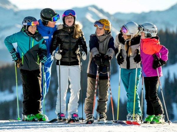 A group of six people in winter clothing and ski gear stands together on a snow-covered mountain, with snowy trees and mountains in the background.