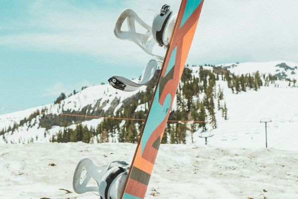 A colorful snowboard standing upright in snowy terrain with mountains and trees in the background under a partly cloudy sky.