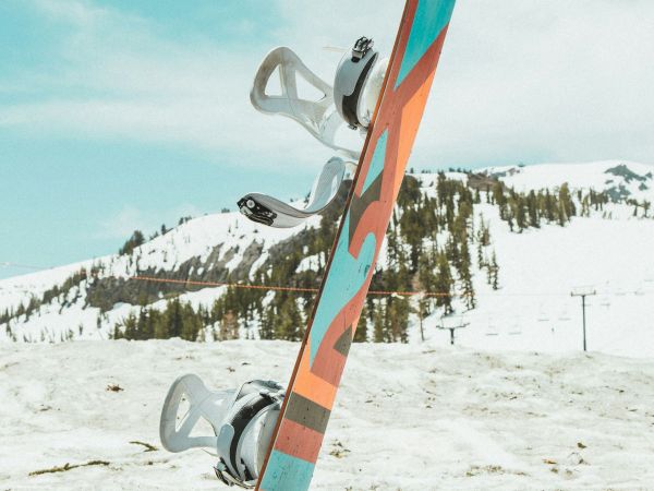 A colorful snowboard standing upright in snowy terrain with mountains and trees in the background under a partly cloudy sky.