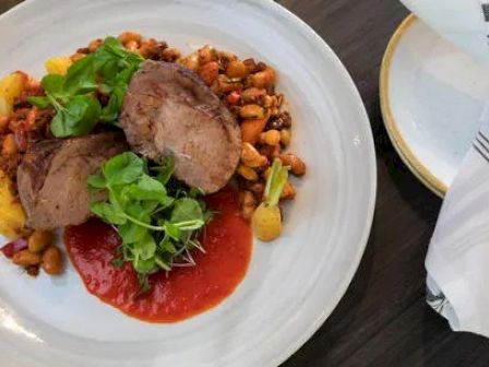 A plate with slices of meat on a bed of beans, sauce, and greens. A folded napkin and empty plate are beside it on a dark table.