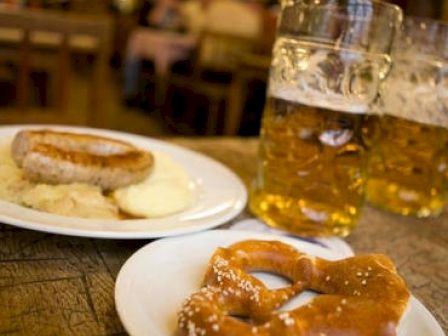 A table with a plate of sausages and mashed potatoes, a plate with a pretzel, and two large mugs of beer in a restaurant setting.