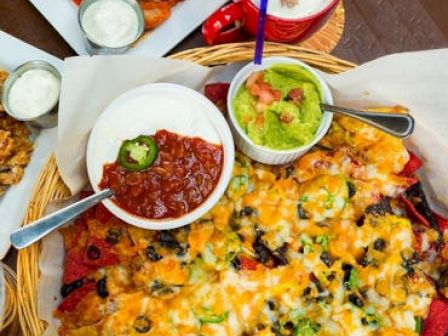 The image shows a basket of nachos topped with melted cheese and served with side dips of guacamole, salsa with a jalapeño, and sour cream.