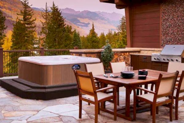 The image shows an outdoor patio with a hot tub, dining table, chairs, and a grill, surrounded by a scenic mountainous forest landscape.