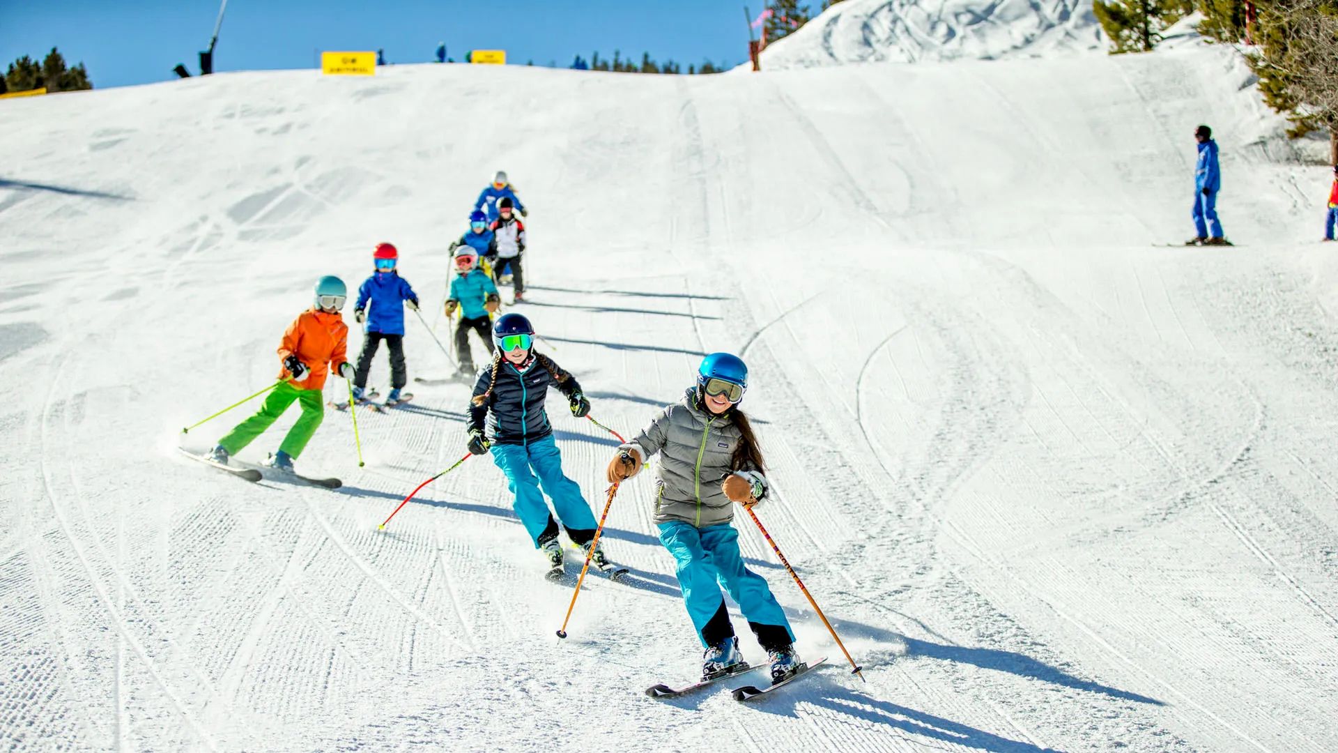 A group of children, wearing colorful winter gear, skiing down a snowy slope with a clear blue sky in the background.