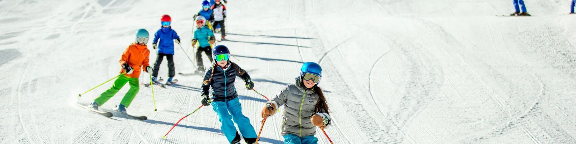 A group of children, wearing colorful winter gear, skiing down a snowy slope with a clear blue sky in the background.