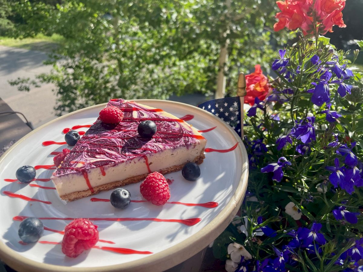 A slice of berry cheesecake garnished with raspberries and blueberries on a plate, set outside next to vibrant flowers in the sunlight.