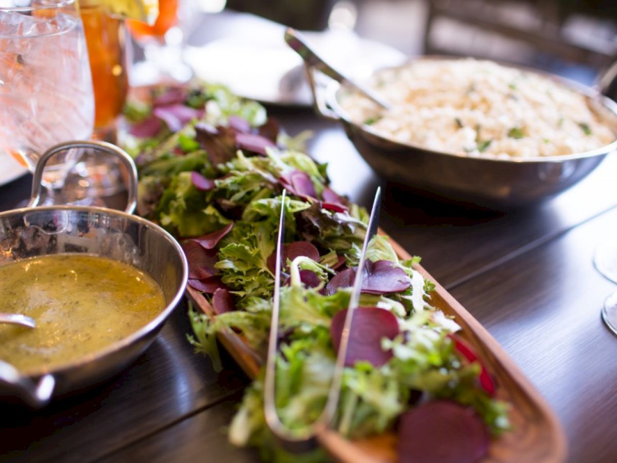 A fresh salad with greens and beets, dressing in a bowl, rice dish, and drinks on a table.