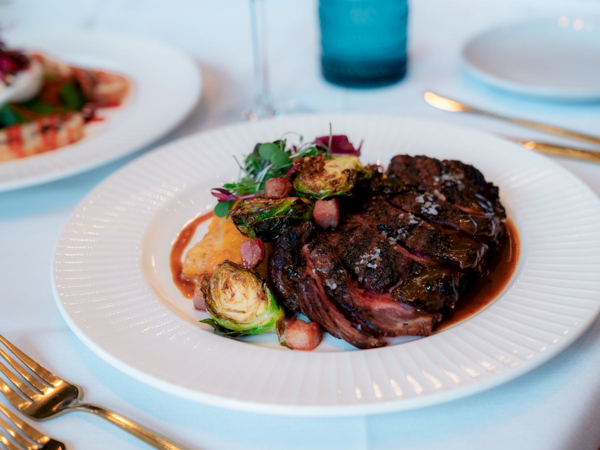 A dish featuring a cooked steak, Brussels sprouts, and garnish on a white plate, set on a table with gold cutlery and a blue glass.