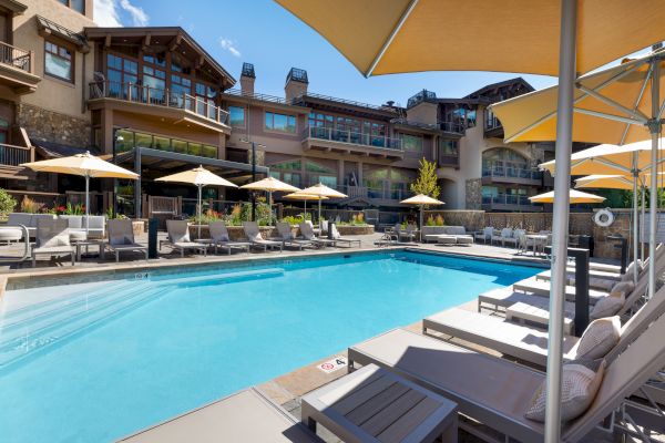 The image shows a luxurious outdoor pool area with numerous lounge chairs and umbrellas, surrounded by a large building with multiple windows.