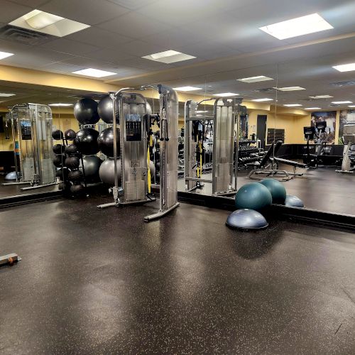 A gym with equipment like weights, exercise balls, and machines, mirrored walls and rubber flooring, under bright ceiling lights.