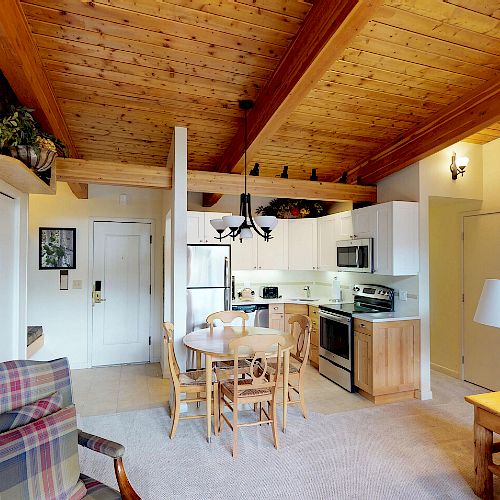 The image shows a cozy living space with wooden ceilings, a small dining table, a kitchen area, a colorful wall painting, and some comfortable furniture.