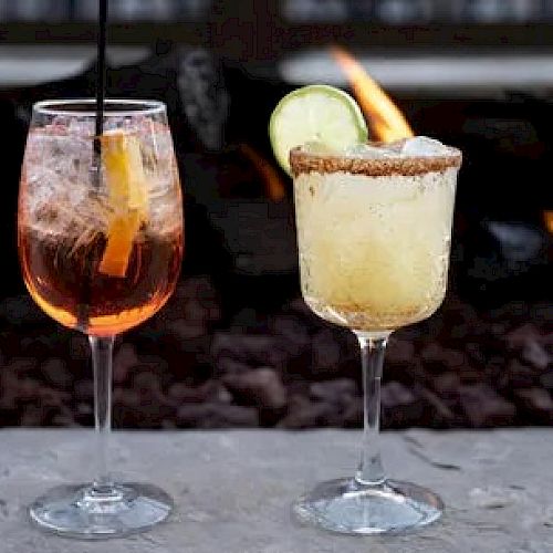 Two cocktails are placed on a surface in front of a lit fire pit, one with a straw and one garnished with a lime and a rim of salt.