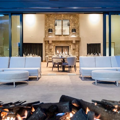 The image shows a modern outdoor seating area with white couches and a fire pit, in front of a stone fireplace in a large, well-lit room.