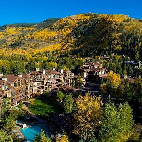 A scenic view of a mountain resort area with lush forests, autumn foliage, and various buildings, including a pool, nestled in a valley at daytime.