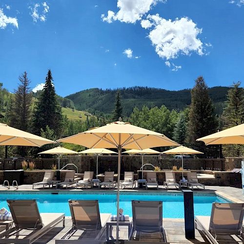 A picturesque swimming pool area with lounge chairs and umbrellas surrounded by a mountainous landscape under a blue sky with scattered clouds.