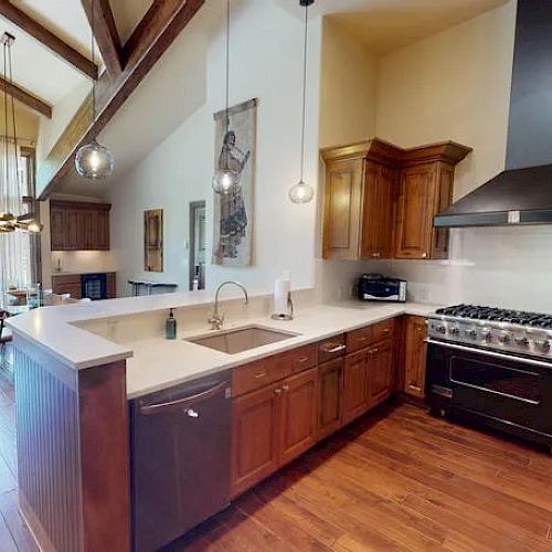 A modern kitchen with wooden cabinets, large island, stainless steel sink, stove with vent hood, and pendant lighting. Dining area in the background.