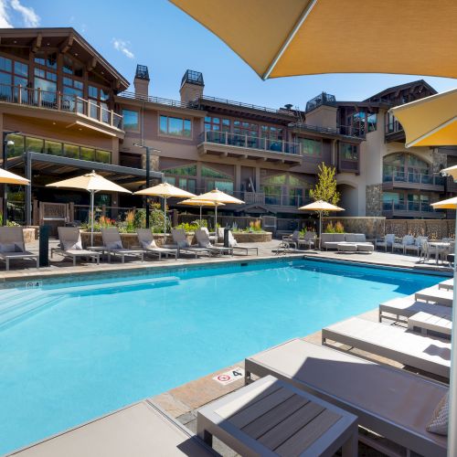 A serene outdoor pool area with lounge chairs, yellow umbrellas, modern architecture in the background, and a clear blue sky.