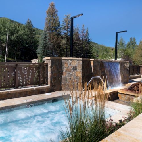 Outdoor spa with hot tubs, stone waterfall feature, surrounded by fence and lush green trees under a clear blue sky.