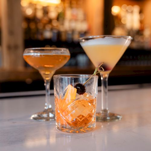 Three cocktails on a bar counter, each in different glassware, with a blurred background of a well-stocked bar.