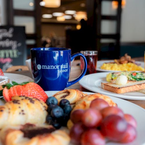 A breakfast spread with pastries, fruits, toast with avocado, juice, and a blue mug, placed on a table.