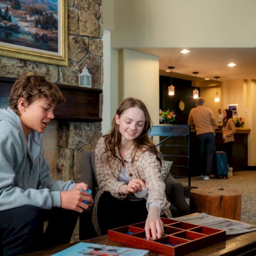 Two people are playing a board game in a cozy living room with artwork, while two others are at a reception desk in the background.