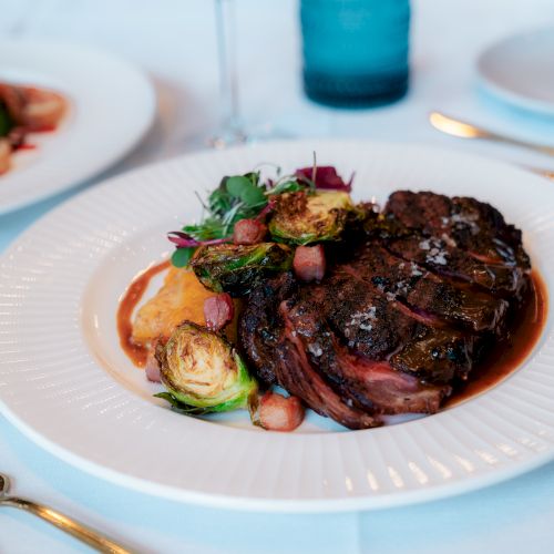 A plated gourmet meal featuring a cooked steak, Brussels sprouts, and garnish, with golden cutlery on a white tablecloth in the background.