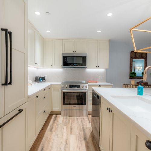 A modern kitchen with white cabinetry, a central island with a sink and faucet, stainless steel appliances, and wooden flooring is visible.