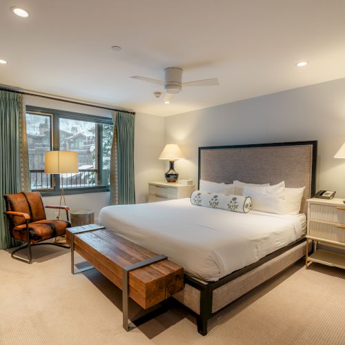 A neatly arranged bedroom with a large bed, two nightstands with lamps, a wooden bench, an armchair, a window with green curtains, and a ceiling fan.