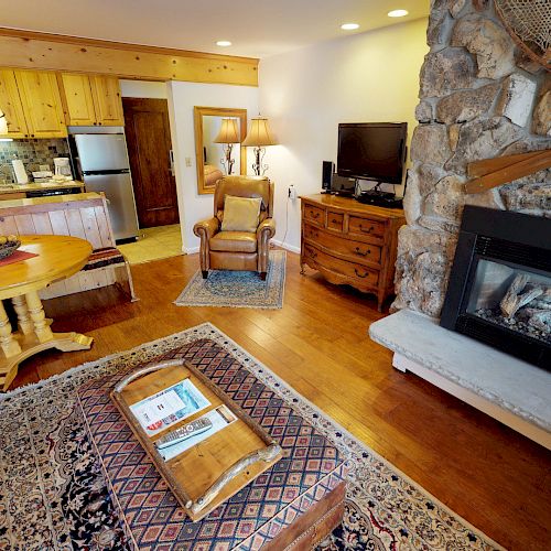 A cozy living room with a stone fireplace, couch, armchair, wooden furniture, and an open kitchen in the background.