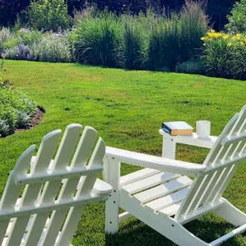 A serene garden scene featuring two white wooden chairs facing lush greenery, with a small table holding a book and a mug, ending the sentence.