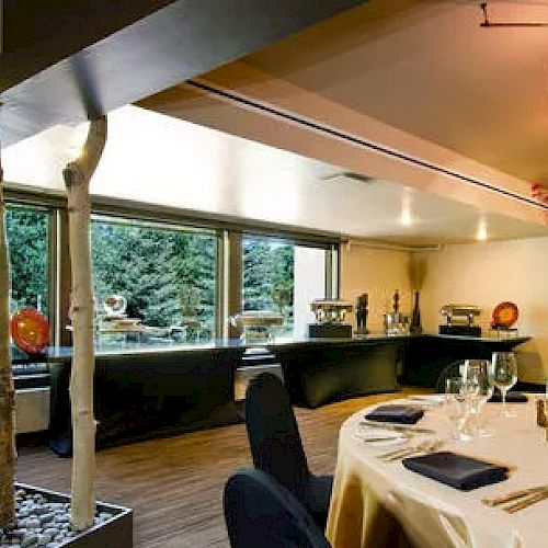 The image shows a modern dining area set for an event, with elegant table settings, tall decorative elements, and a buffet table in the background.