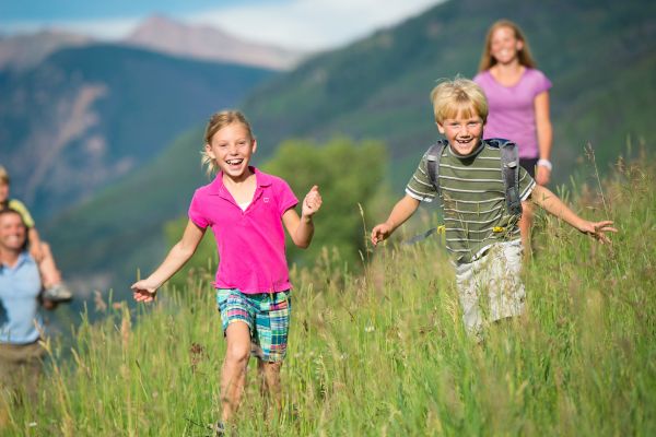 Two children running happily through a grassy field with mountains in the background on a sunny day.