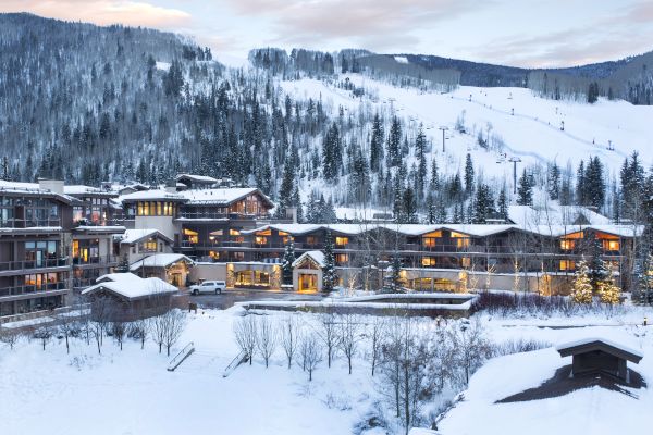 The image shows a snowy mountain resort with multiple buildings surrounded by trees and hills, under a clear sky at dusk.
