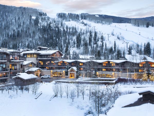 The image shows a snowy mountain resort with multiple buildings surrounded by trees and hills, under a clear sky at dusk.