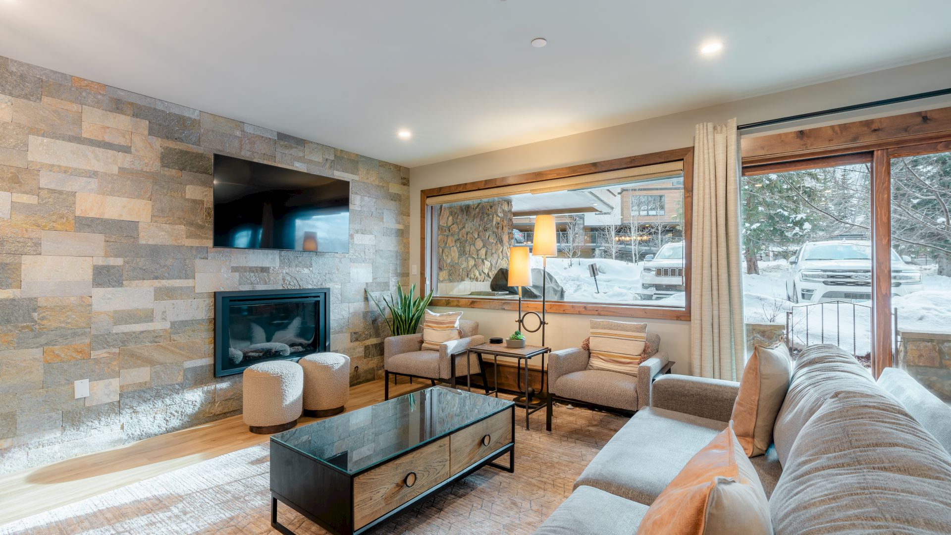 A cozy living room with modern furnishings, stone accent wall, fireplace, large windows, and outside snowy view creating a warm atmosphere.