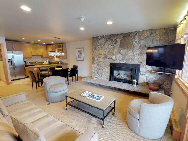 A cozy living room with a stone fireplace, wall-mounted TV, beige furniture, coffee table, and a connected kitchen area.