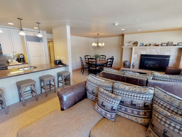 This image shows a cozy living room and kitchen area with a bar, stools, a sectional sofa, a dining table with chairs, and a fireplace.
