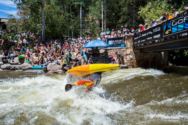 A kayaker performs a trick in a river while a large crowd watches from the riverside and a bridge on a sunny day, amid a vibrant atmosphere.