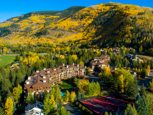 Aerial view of a mountain resort area with buildings surrounded by trees in autumn colors, set against a background of hills and blue sky.
