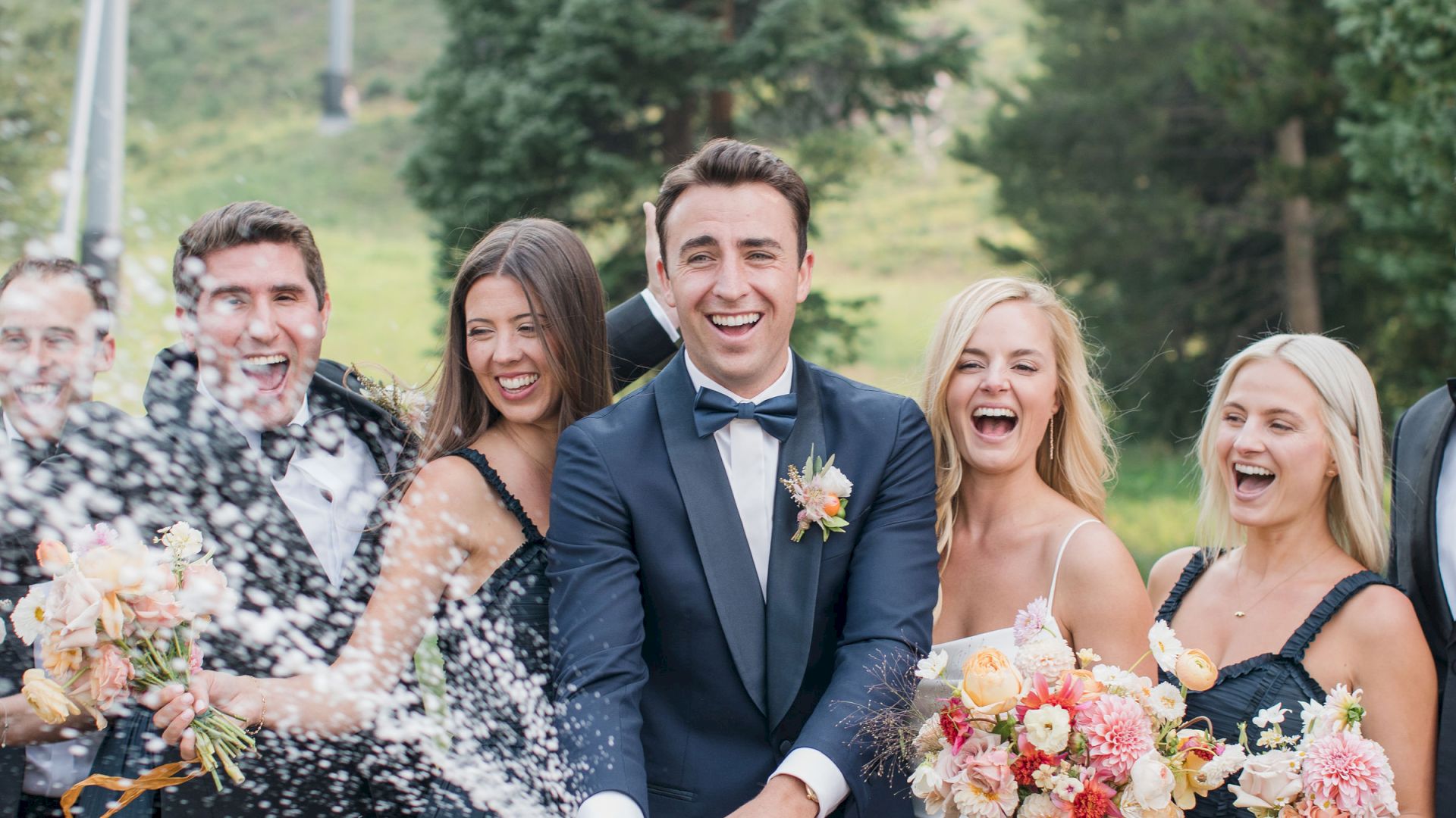 A wedding party is celebrating outdoors. The groom is popping a bottle of champagne, and everyone is smiling in joyful celebration.