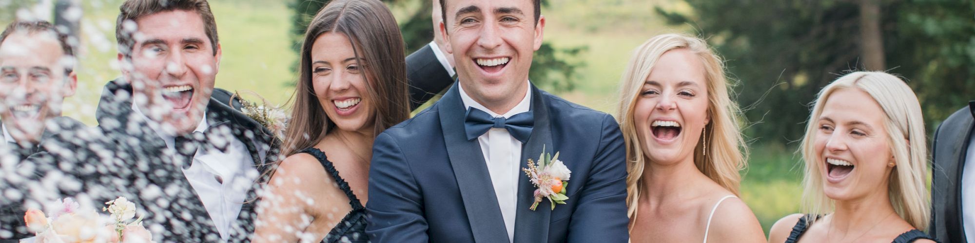 A wedding party is celebrating outdoors. The groom is popping a bottle of champagne, and everyone is smiling in joyful celebration.